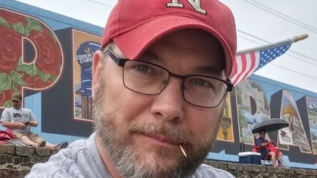 Keith smiling in a red Nebraska cap and glasses, with an American flag behind him