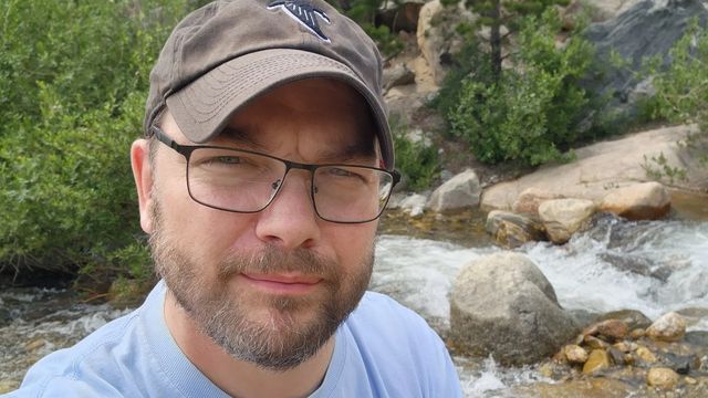 Keith in a brown cap and glasses, taking a selfie beside a rocky mountain stream