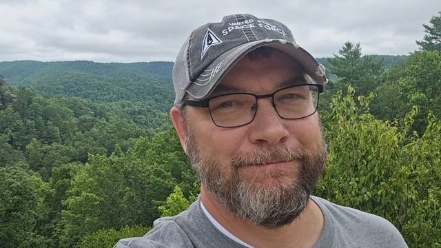 Keith in a gray cap and glasses, taking a selfie at a scenic overlook above green forested hills
